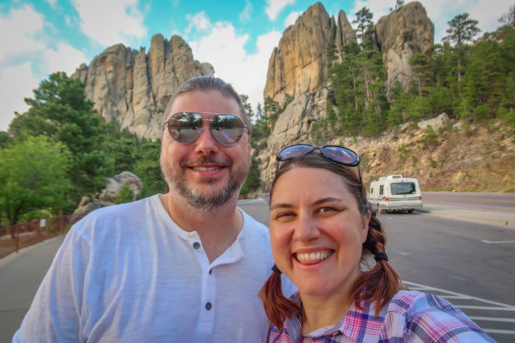Photo of the author with her husband Daniel in front of Mount Rushmore as viewed from South Dakota Highway 244. Only a portion of Washington's head is visible from this angle. Our Winnebago Rialta motorhome is parked in the background.
