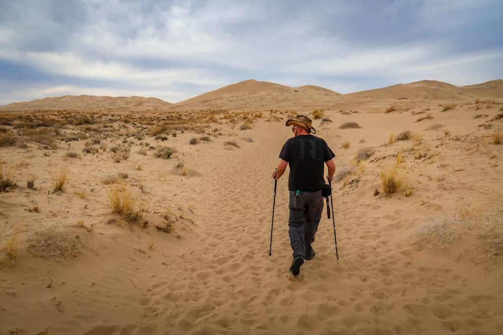 Daniel hikes the Kelso Dunes Trail, using hiking poles and wearing a broad straw hat