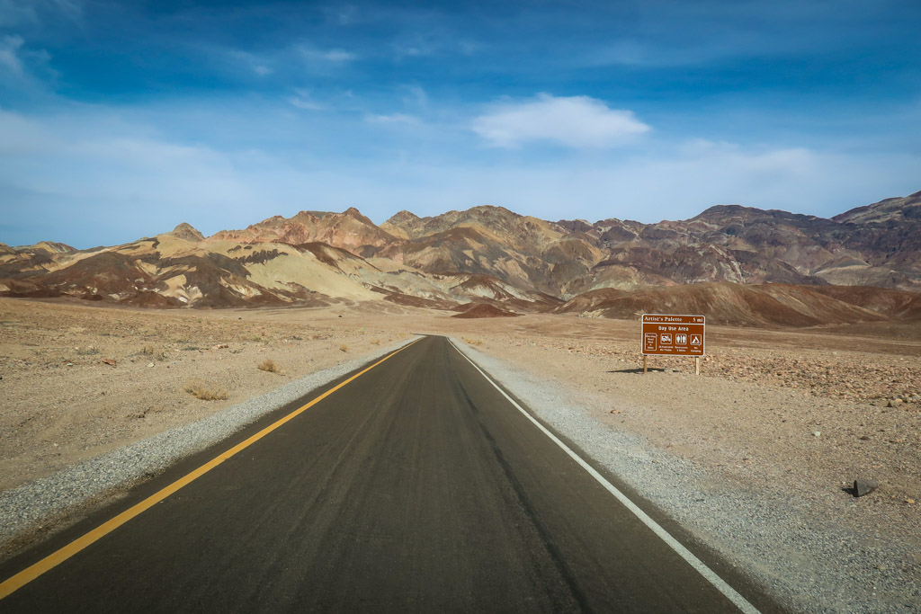 The road in Death Valley stretches ahead with colorful hills in the distance streaked with various vivid shades of brown and white and red. These are the Artist's Palette.