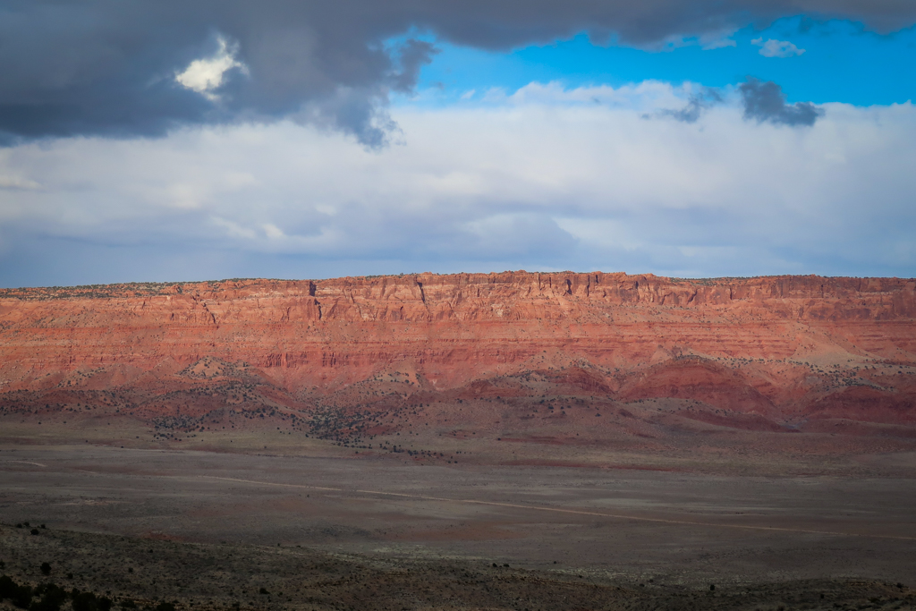 A towering wall of cliffs that rise dramatically from a plateau floor. The cliffs are formed of various shades of red and can be seen when driving the Vermillion Cliffs Scenic Highway.