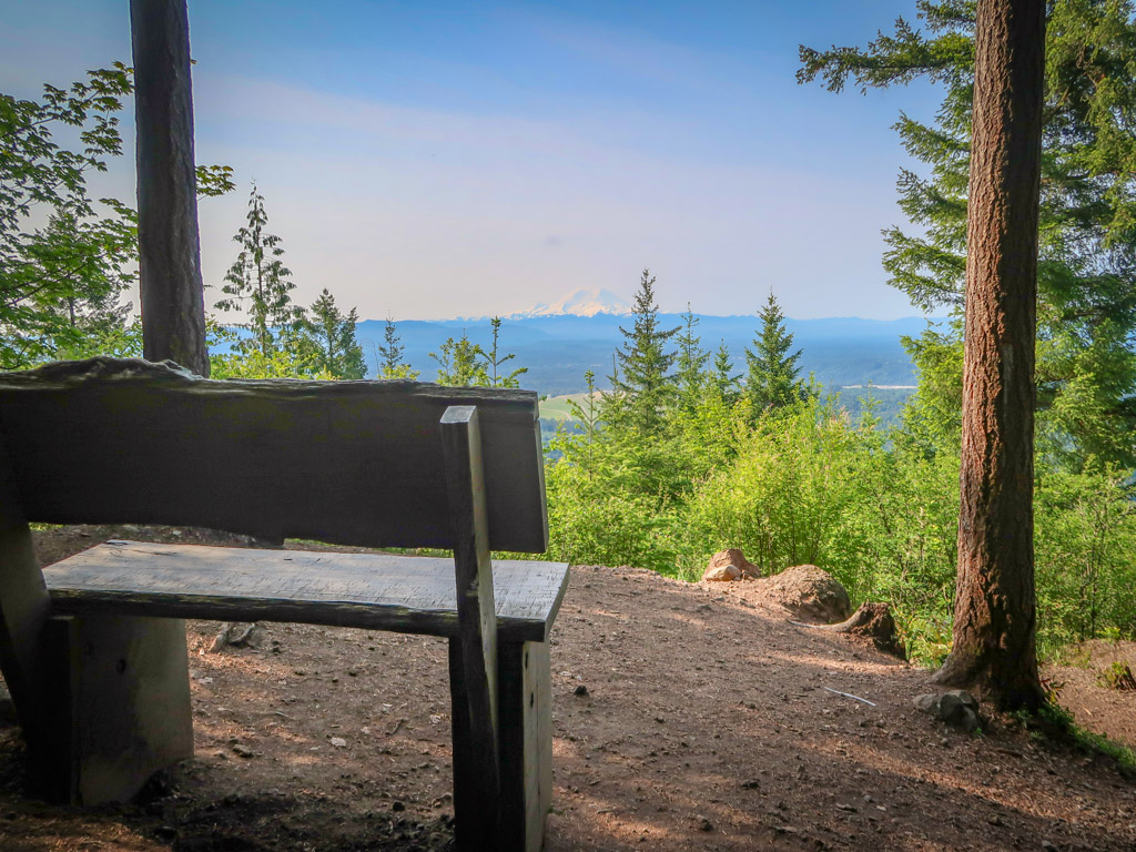 A bench at Debbie's View sits facing a vista with Mt. Rainier poking above the clouds in the distance