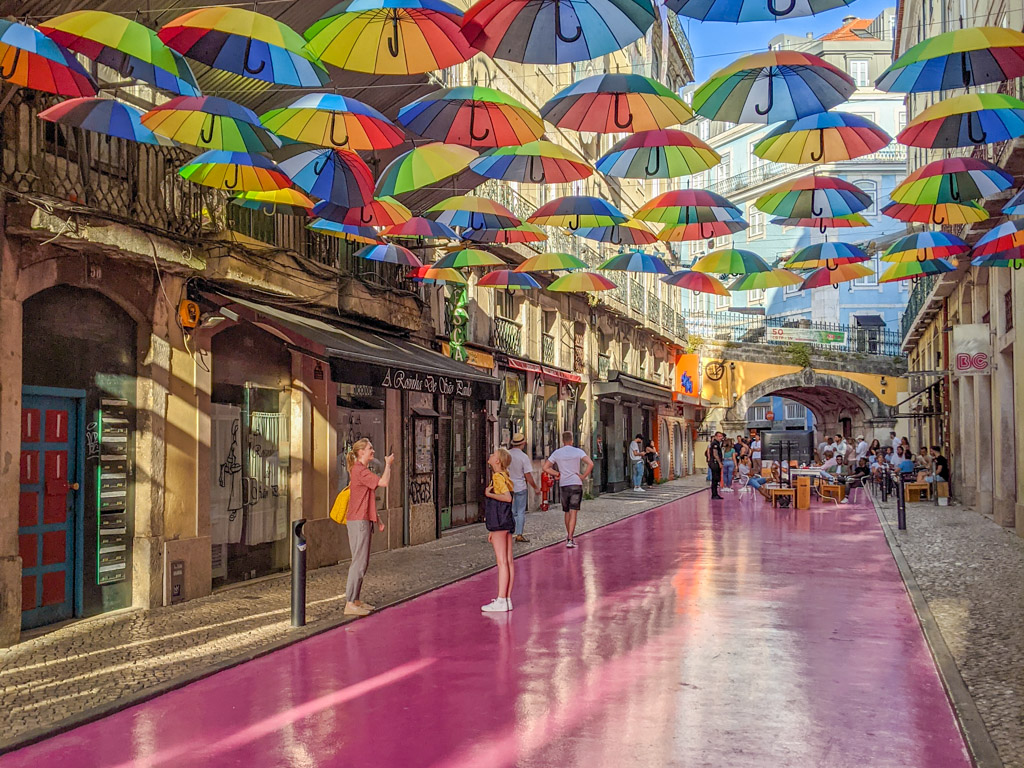 Brightly-colored rainbow-striped umbrellas are strung across a street painted bright street an an alley lined with shops and cafes.