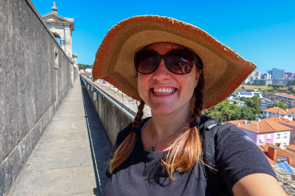 Photo of the author with a broad straw sun hat with braids and sunglasses standing atop the Lisbon Aqueduct