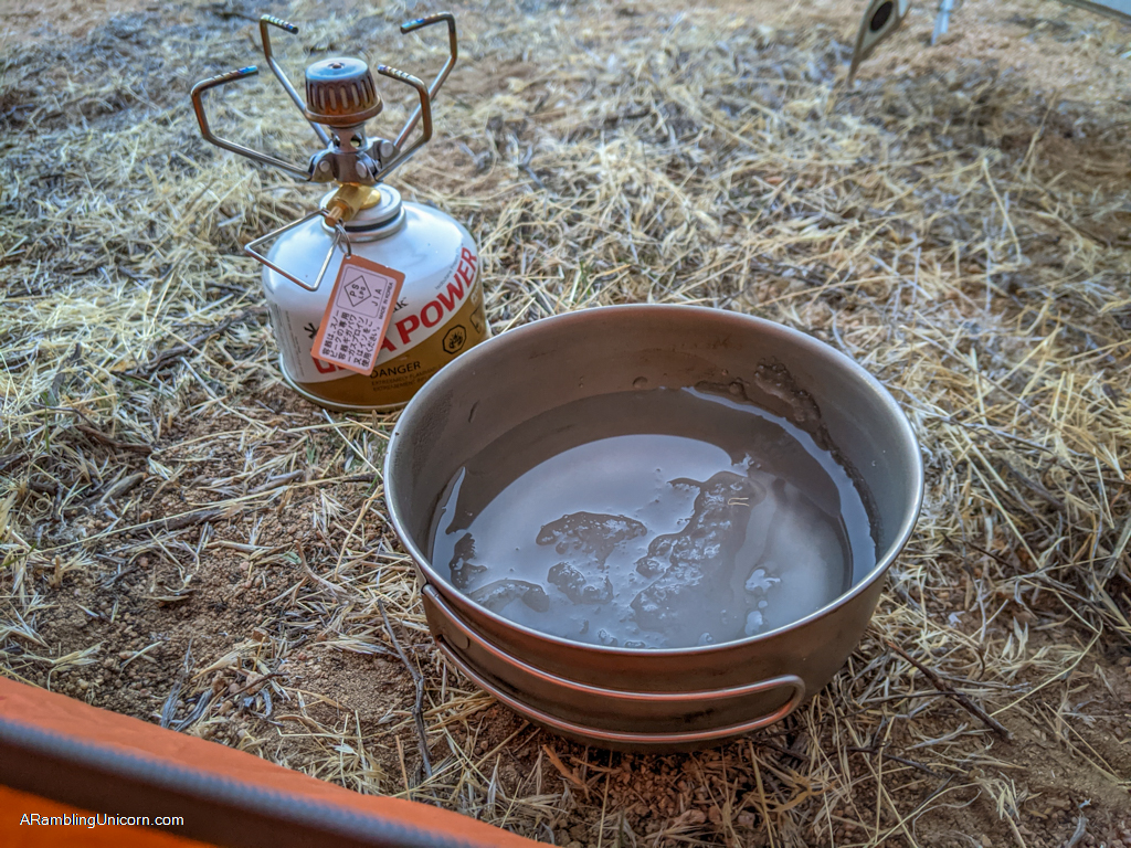Cooking pot with partially frozen slushy water in it next to a backpacking stove affixed to a small propane canister in the tent vestibule.
