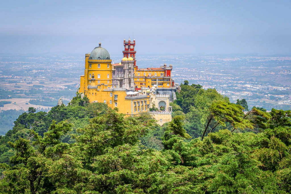 A distant view of Pena Palace perched high on a hill overlooking Sintra. The place is painted a distinctive bright yellow and red color scheme.