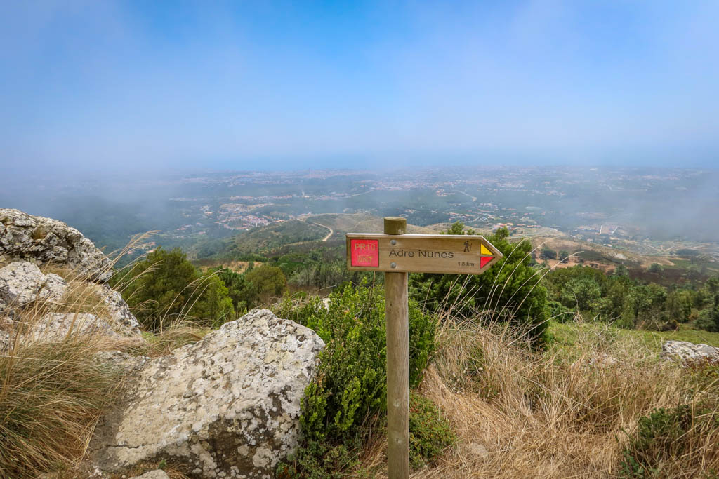 A hiking signpost stands on an overlook. The marker reads "PR10. Andre Nunes 1,8 km" 