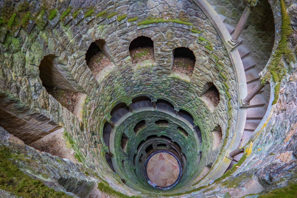 A spiral staircase leads down an inverted stone tower that is covered in green moss