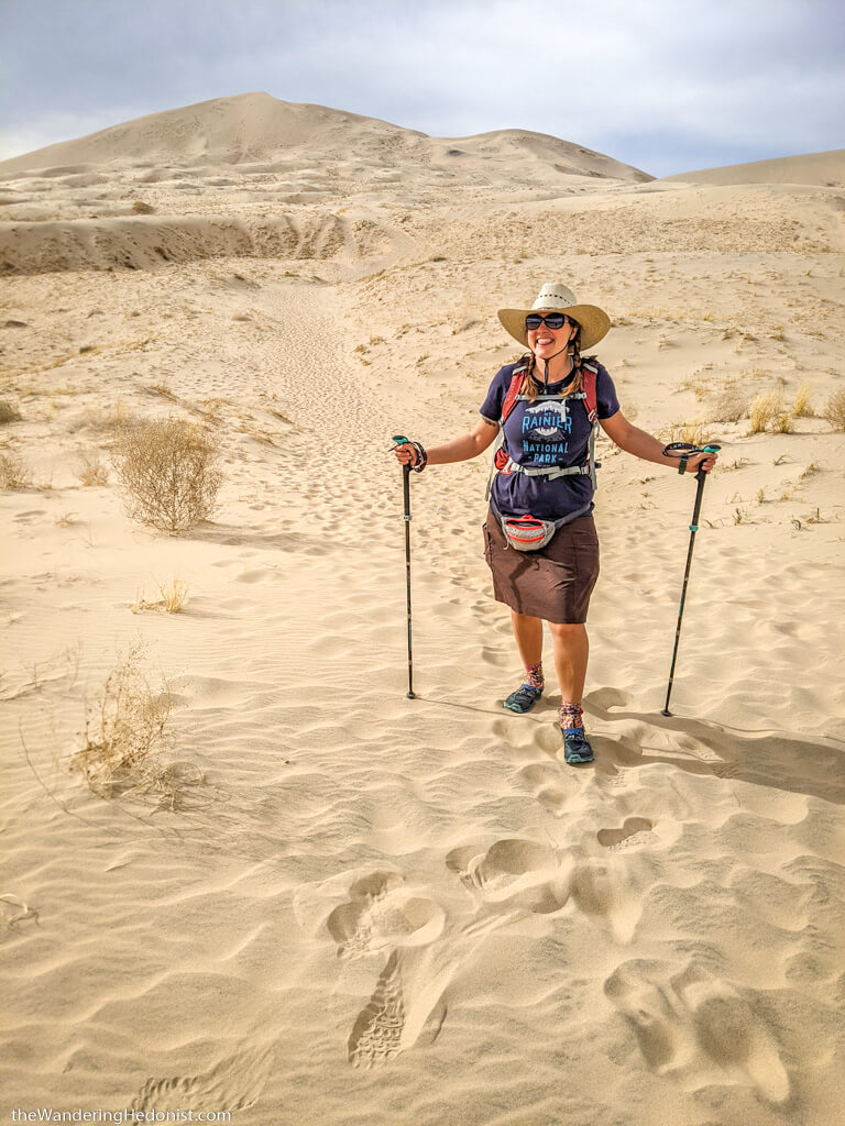 the author poses in front of the Kelso Dunes with hiking poles and broad straw hat