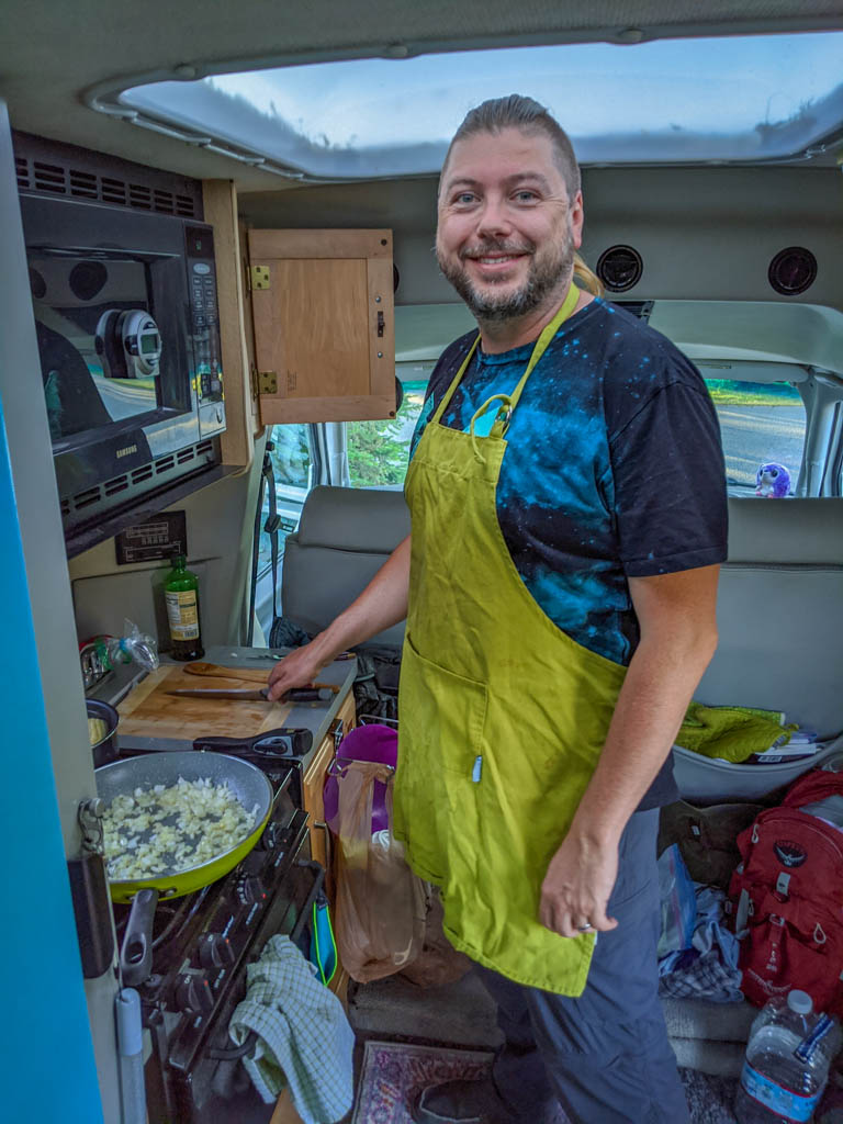 Daniel cooks onions in a skillet inside of the motorhome while wearing a bright green apron.