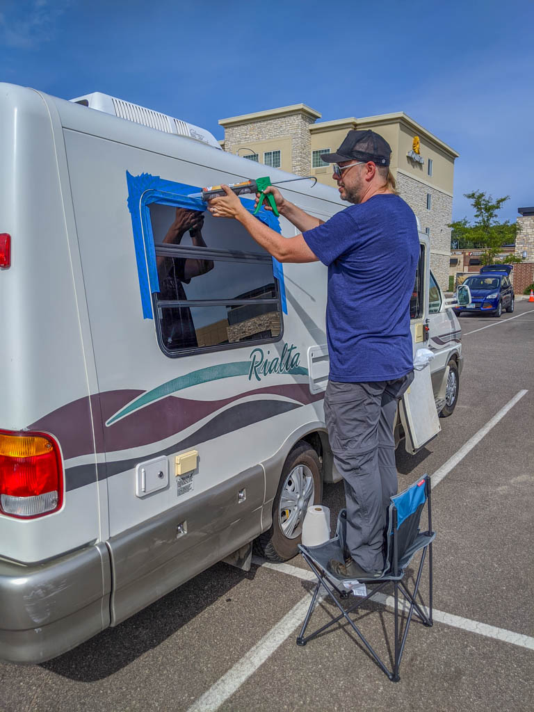 Daniel stands on a chair in a hotel parking lot to caulk around the windows to prevent leaks