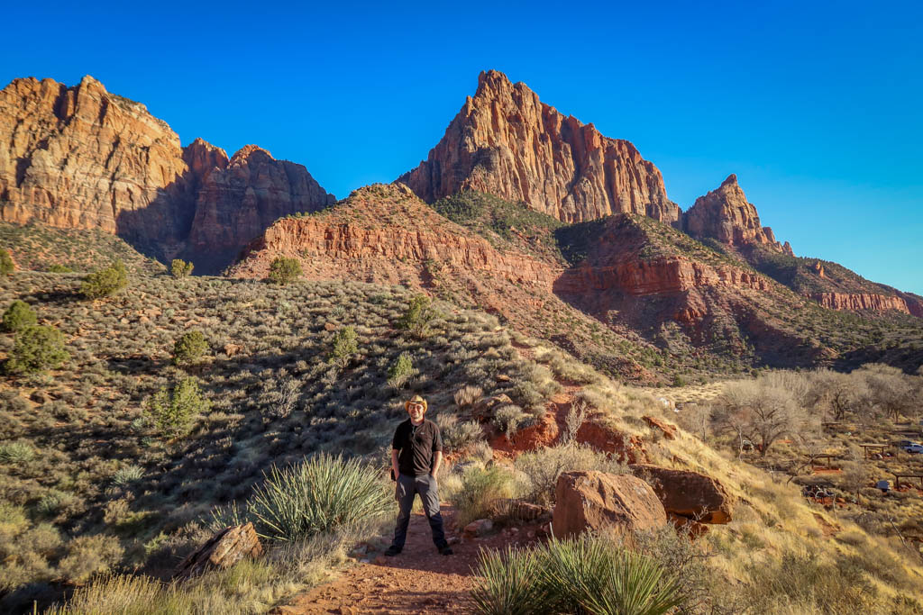 Daniel stands on a hiking path with the tall jagged red walls of Zion Canyon framed behind him.