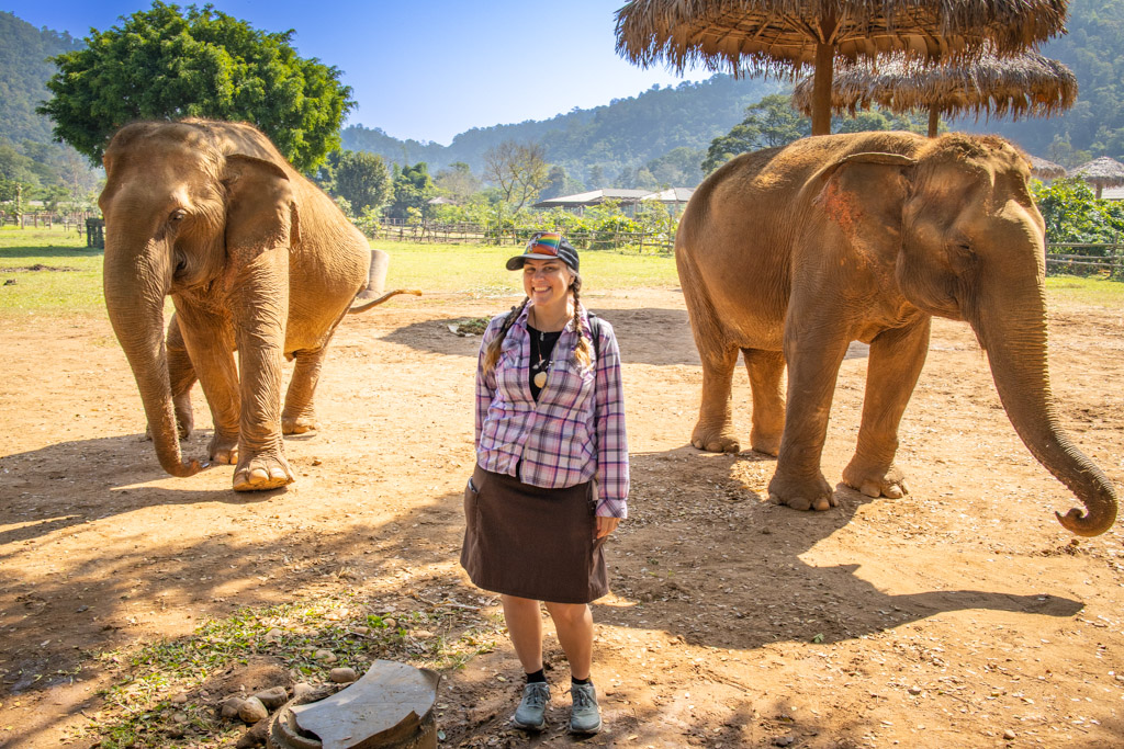 The author is pictured with 2 Asian elephants in the background at Elephant Nature Park, known as one of the best Thailand elephant sanctuary